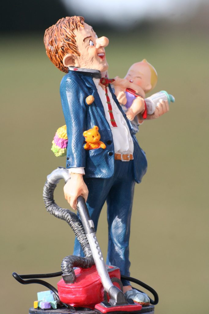 Clay figurine of a man holding a baby and the hoze of a vauum cleaner, wearing a suit with baby stuff sticking out of every pocket